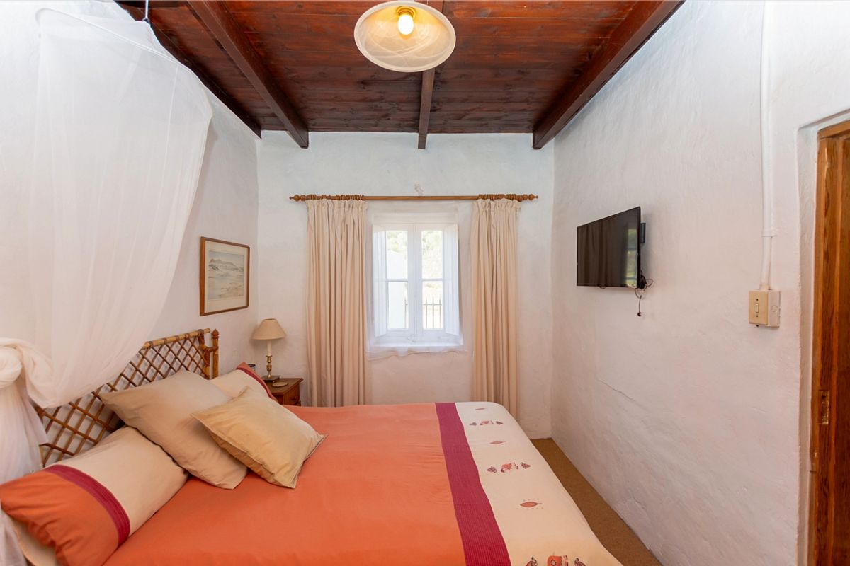 A cozy bedroom featuring rustic exposed timber ceiling beams, white textured walls, and a central window with natural light.