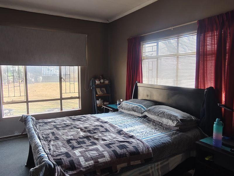 A bedroom featuring aluminium window frames with security bars, roller blinds, and curtain tracks, providing natural light and garden views.