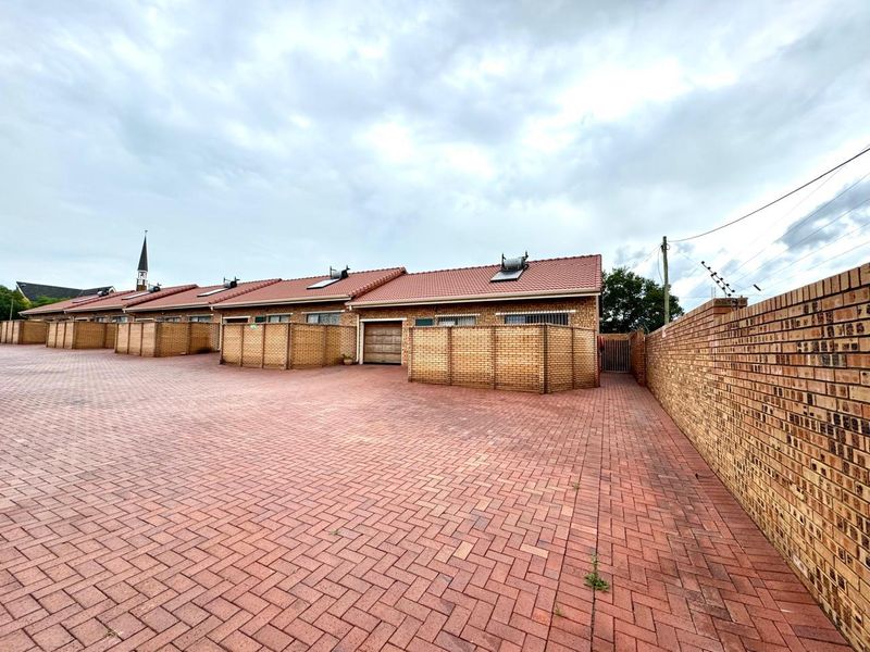 Exterior view of a residential apartment complex featuring brick paved driveway, perimeter walling, and tiled roof structures with solar geysers.