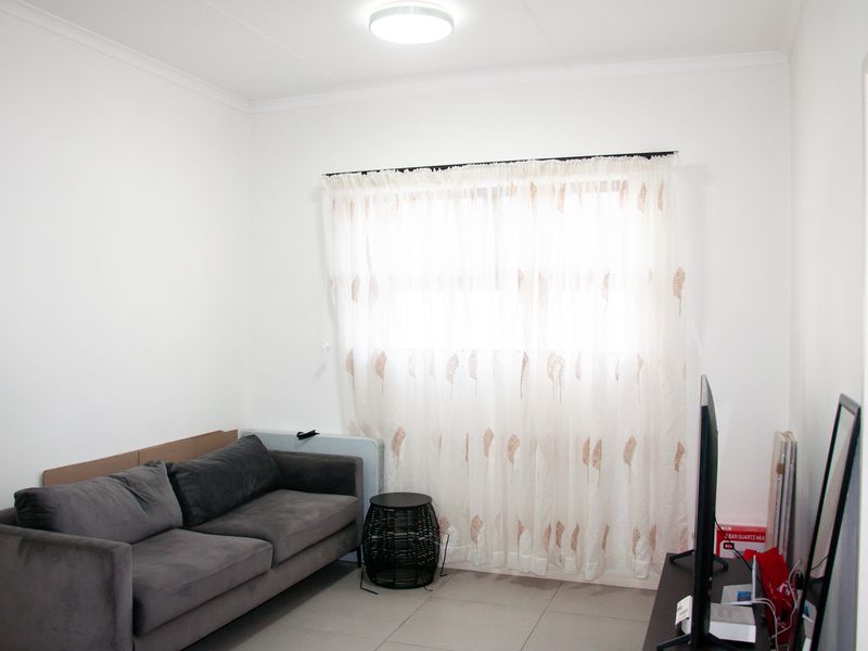 A simple living room featuring neutral tiled flooring, white walls, and a window fitted with a curtain rod.