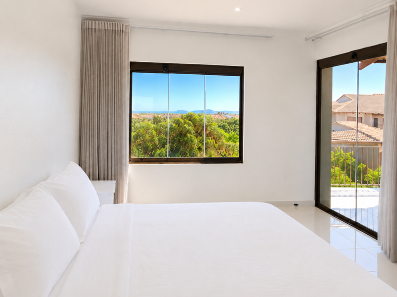 A bright, modern bedroom featuring tiled floors, large sliding glass doors leading to a balcony, and expansive views of the surrounding landscape.