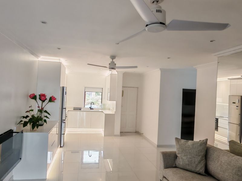 A bright, modern open-plan living and kitchen area featuring high-gloss cabinetry, polished tiled floors, recessed lighting, and a large mirrored feature wall.