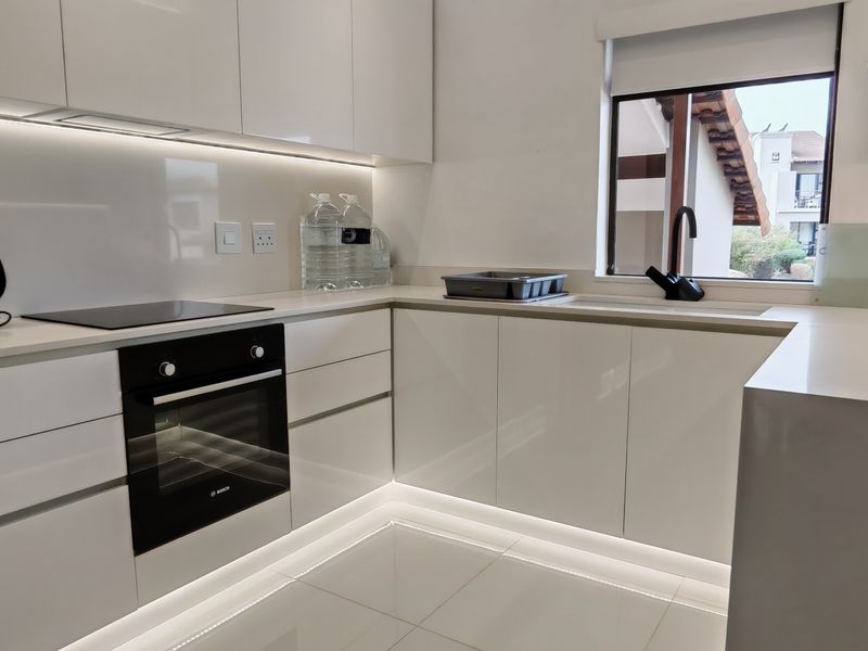Modern, well-lit kitchen featuring sleek white gloss cabinetry, stone countertops, integrated appliances, and contemporary under-cabinet and plinth lighting.