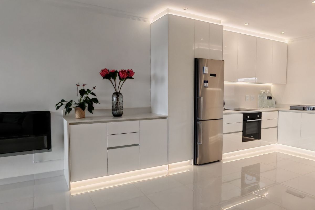 Modern kitchen featuring sleek white cabinetry, integrated under-cabinet lighting, built-in oven and hob, and polished tile flooring, adjacent to a wall-mounted fireplace.