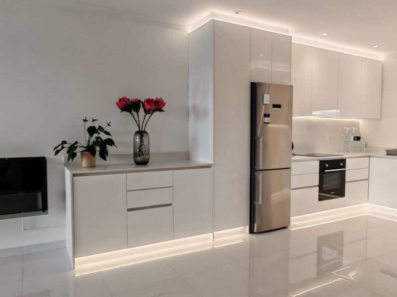 Modern kitchen featuring sleek white cabinetry, integrated under-cabinet lighting, built-in oven and hob, and polished tile flooring, adjacent to a wall-mounted fireplace.