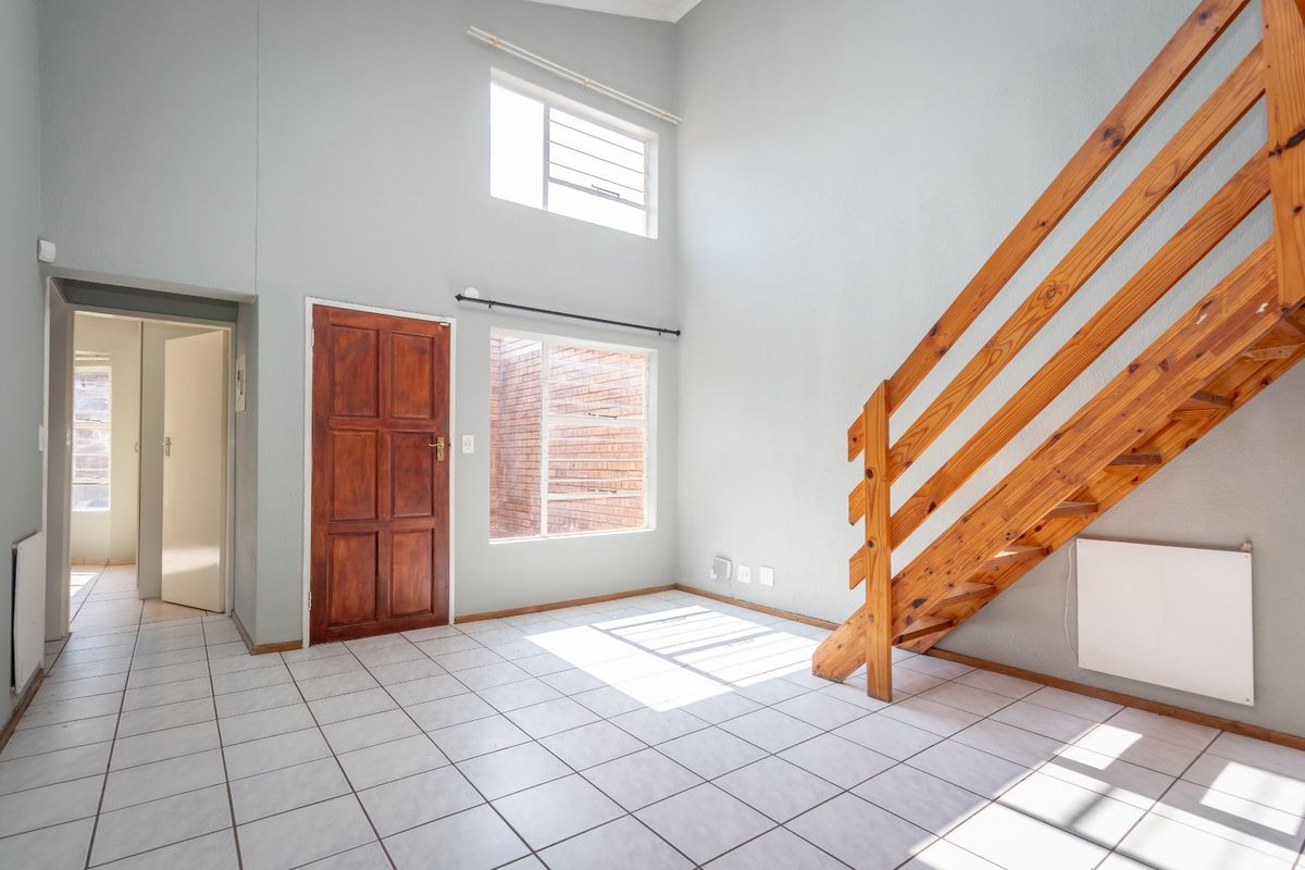 Bright, double-volume living area featuring tiled flooring, a wooden staircase, and natural light from large windows, offering a clean, blank canvas for interior design.