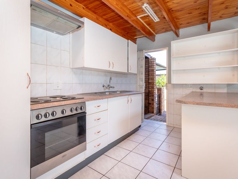 Functional kitchen featuring white cabinetry, laminate countertops, built-in oven with hob, tiled flooring, and exposed timber ceiling beams with direct access to the exterior.
