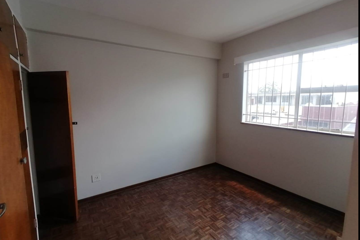 A vacant bedroom featuring classic parquet flooring, a built-in wooden cupboard, and a window fitted with security bars.