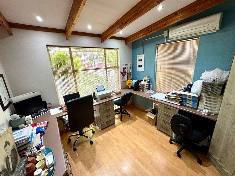 A functional home office featuring exposed timber ceiling beams, built-in L-shaped desk cabinetry, laminate flooring, and a wall-mounted split-system air conditioner.
