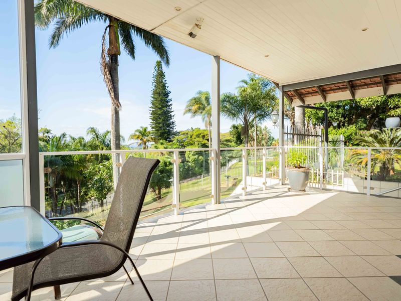 A spacious covered patio featuring tiled flooring and a modern glass balustrade, offering elevated views of the surrounding garden and landscape.