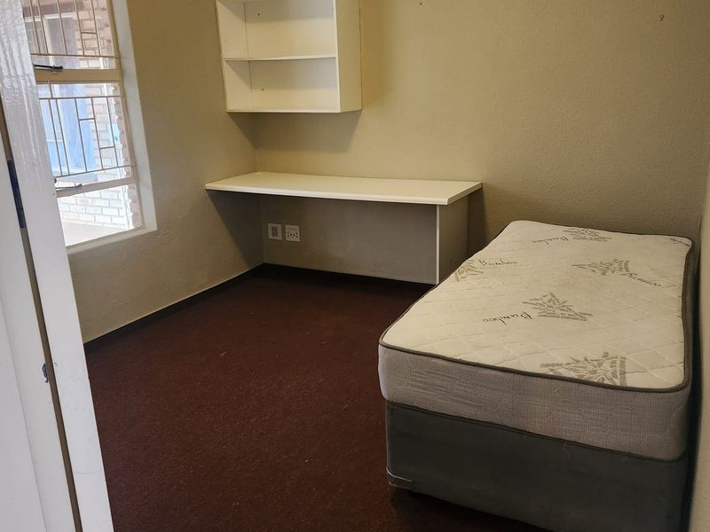 A compact bedroom featuring a built-in desk, wall-mounted shelving, and a window with security bars. The room requires cosmetic updates to improve its appeal.