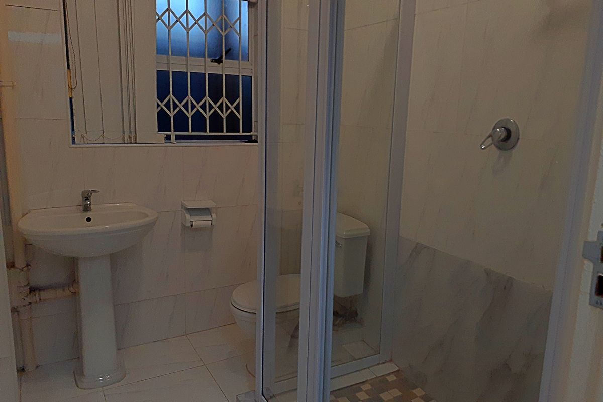 A functional bathroom featuring a pedestal basin, toilet, and a glass-enclosed shower with tiled finishes and security bars on the window.