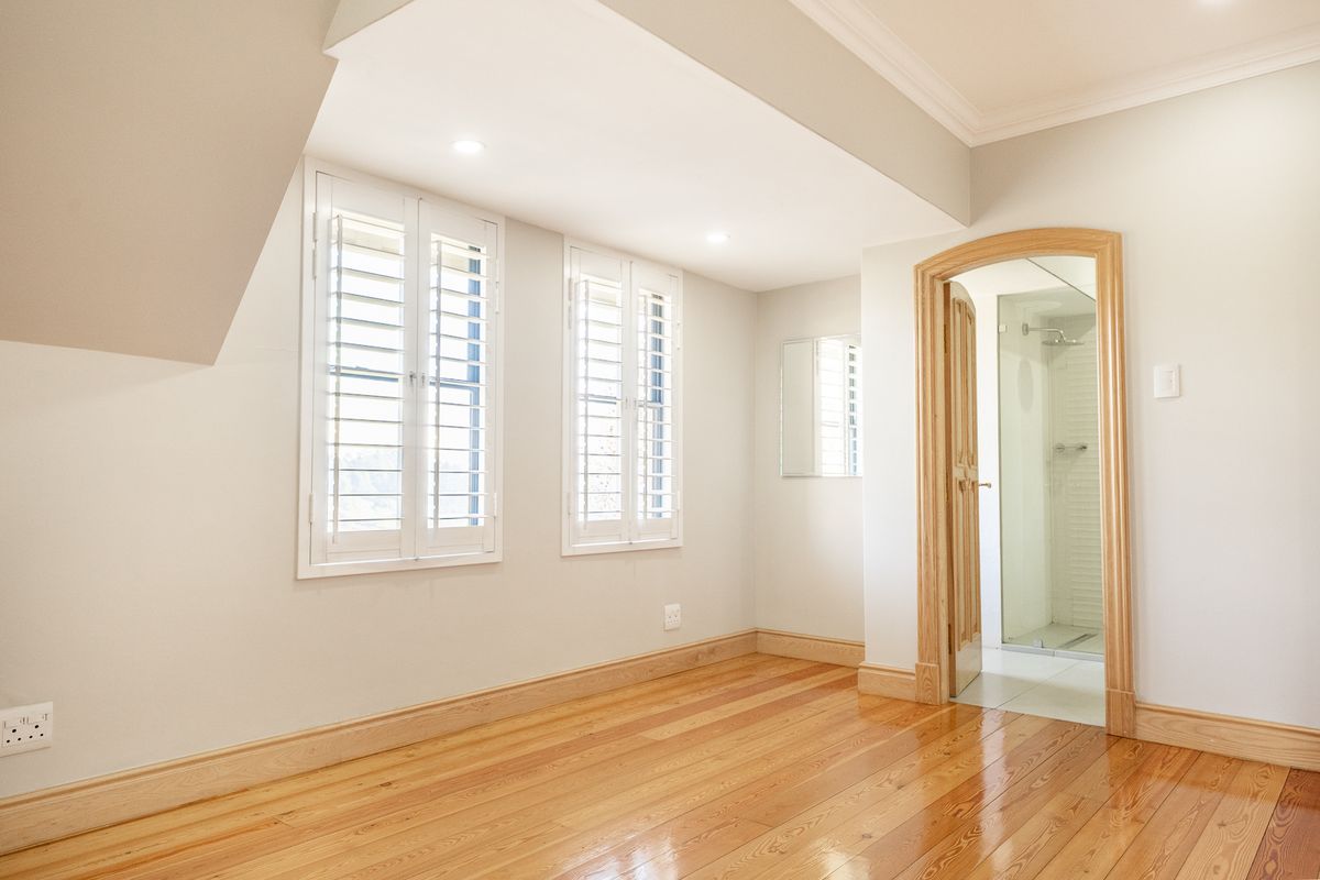 A bright, empty bedroom featuring polished timber flooring, plantation shutters, and direct access to an en-suite bathroom with a glass-enclosed shower.