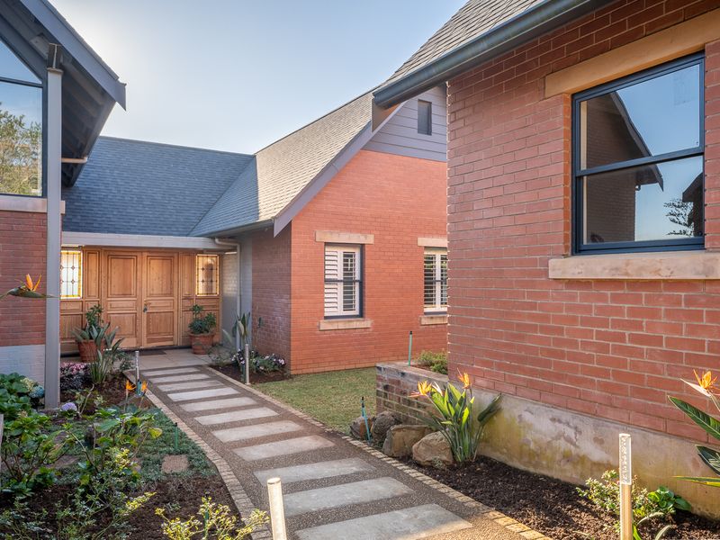 A well-maintained brick residence featuring a stone paved walkway leading to a grand timber entrance, surrounded by established garden beds.
