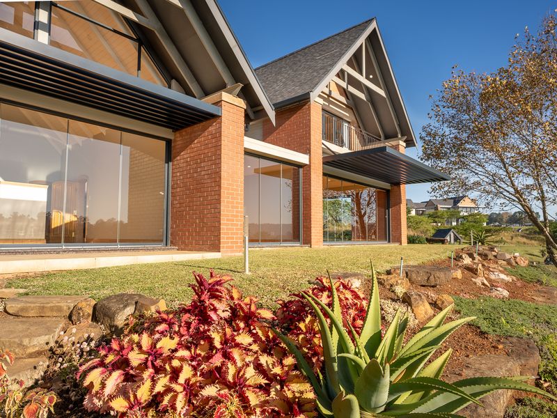 A modern double-story home featuring a red brick facade, expansive glass sliding doors, and a well-maintained landscaped garden with stone retaining walls.