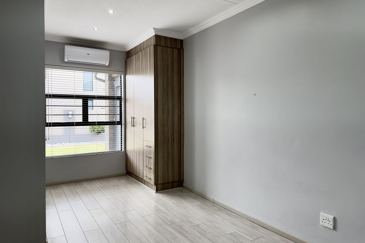 A bright bedroom featuring light wood-look flooring, built-in wooden cabinetry, a wall-mounted air conditioning unit, and large windows with fitted blinds.