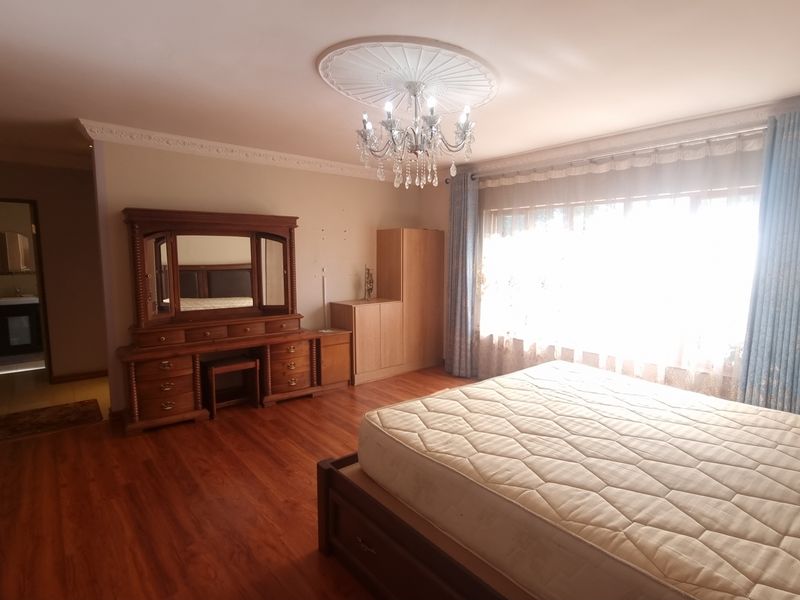 Spacious bedroom featuring laminate flooring, decorative ceiling rose with chandelier, and built-in cupboards, with an en-suite bathroom visible through the doorway.
