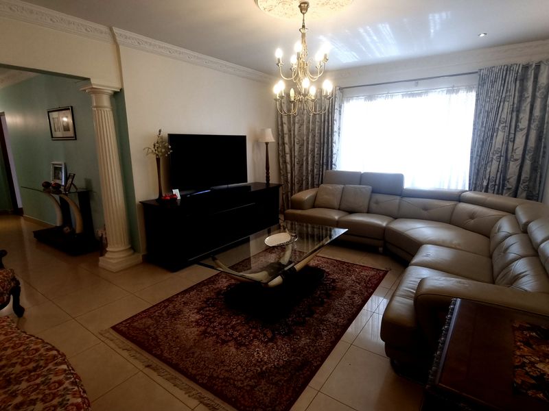 A spacious living room featuring tiled flooring, decorative ceiling cornicing, a classic chandelier, and a structural column leading to an adjacent hallway.
