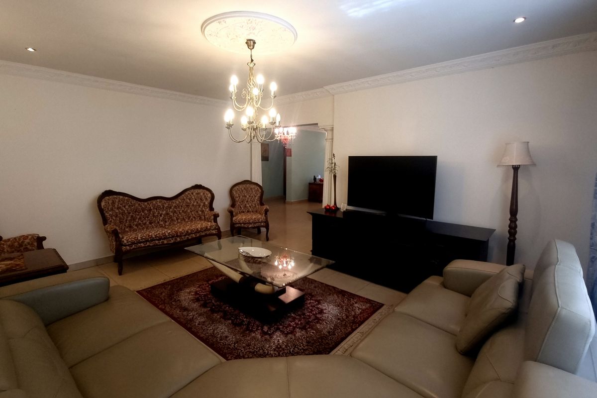 A spacious living room featuring decorative crown molding, a central ceiling rose with a chandelier, and tiled flooring.