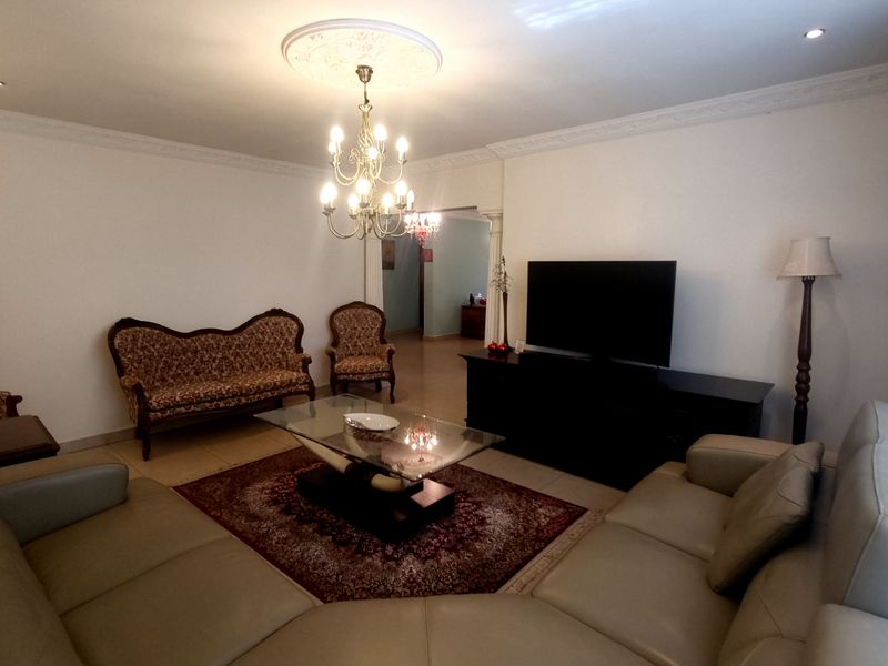 A spacious living room featuring decorative crown molding, a central ceiling rose with a chandelier, and tiled flooring.
