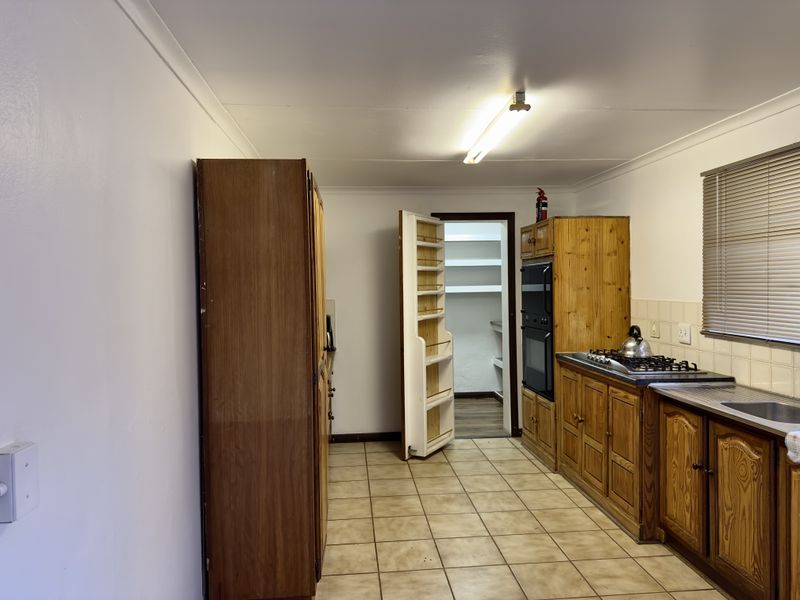 A functional kitchen featuring wooden cabinetry, a gas hob, built-in oven, and an adjoining walk-in pantry with tiled flooring throughout.