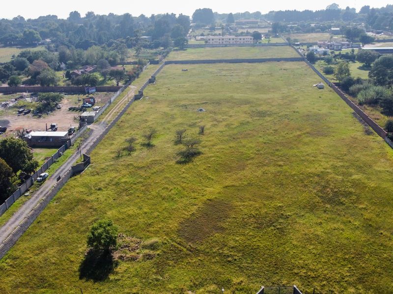 An aerial view of a large, vacant residential plot enclosed by perimeter walls, featuring open grass land and adjacent road access.