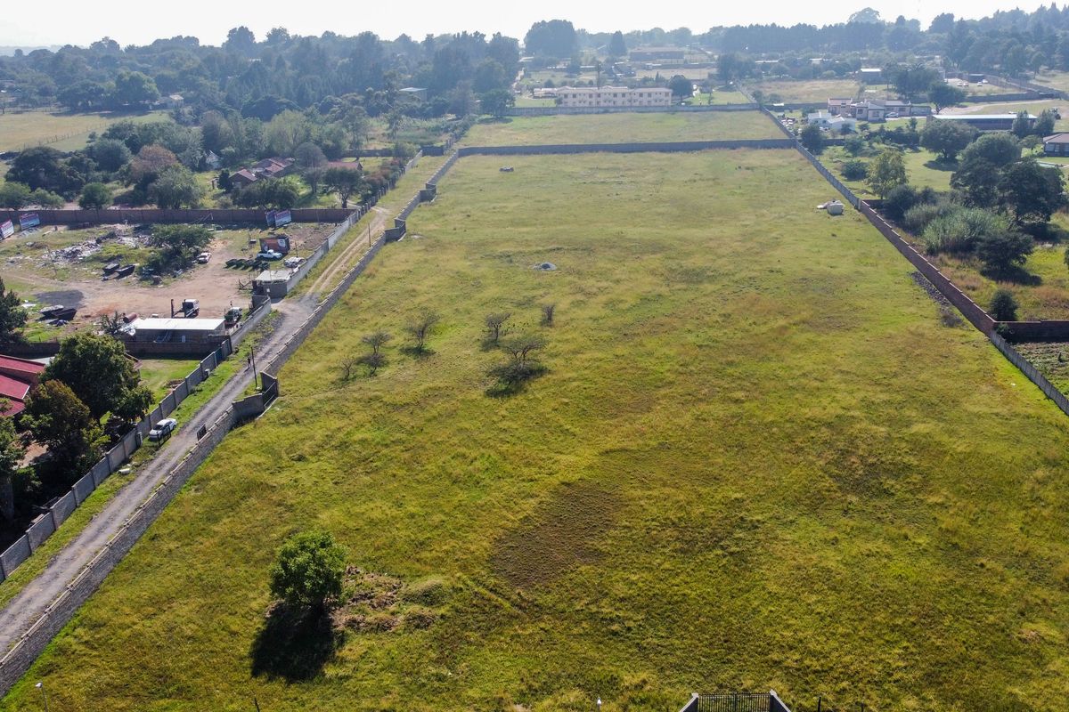 An aerial view of a large, vacant residential plot enclosed by perimeter walls, featuring open grass land and adjacent road access.