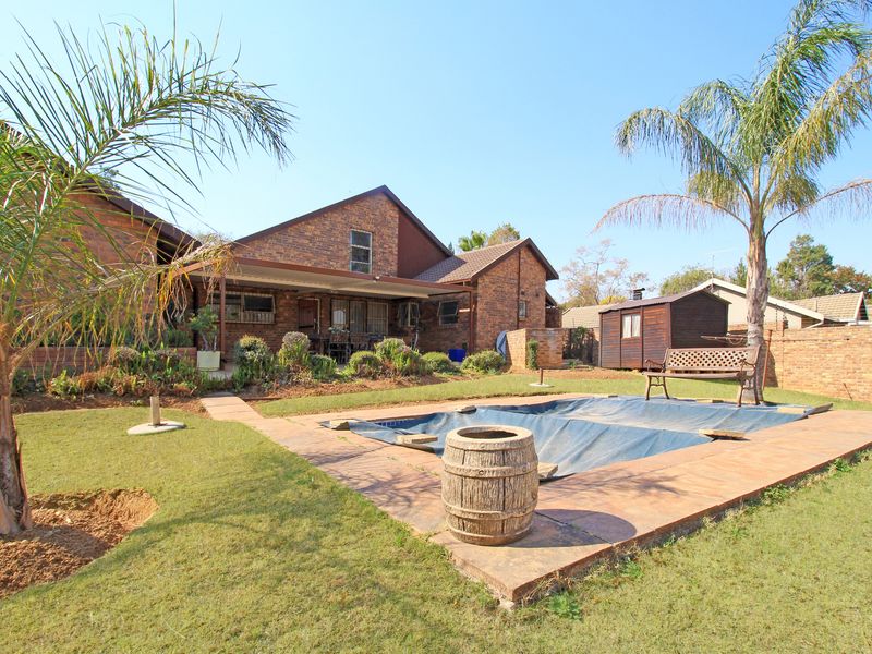 A backyard view featuring a swimming pool with a cover, a covered patio area, and a brick-faced house exterior set within a landscaped garden.