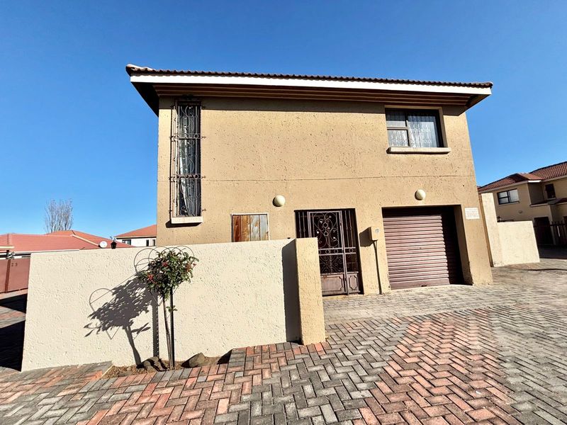 Front elevation of a two-story townhouse featuring a paved driveway, single roller garage door, and secure gated entrance.