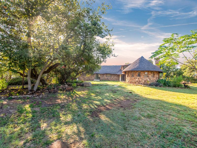 A scenic view of the rear elevation of a stone-clad home featuring a traditional thatched roof, set within a spacious, tree-lined garden.
