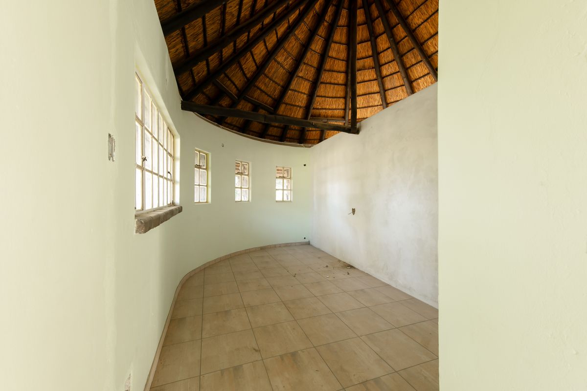 An empty, circular living area featuring a traditional thatched roof with exposed timber rafters, tiled flooring, and multiple steel-framed windows.
