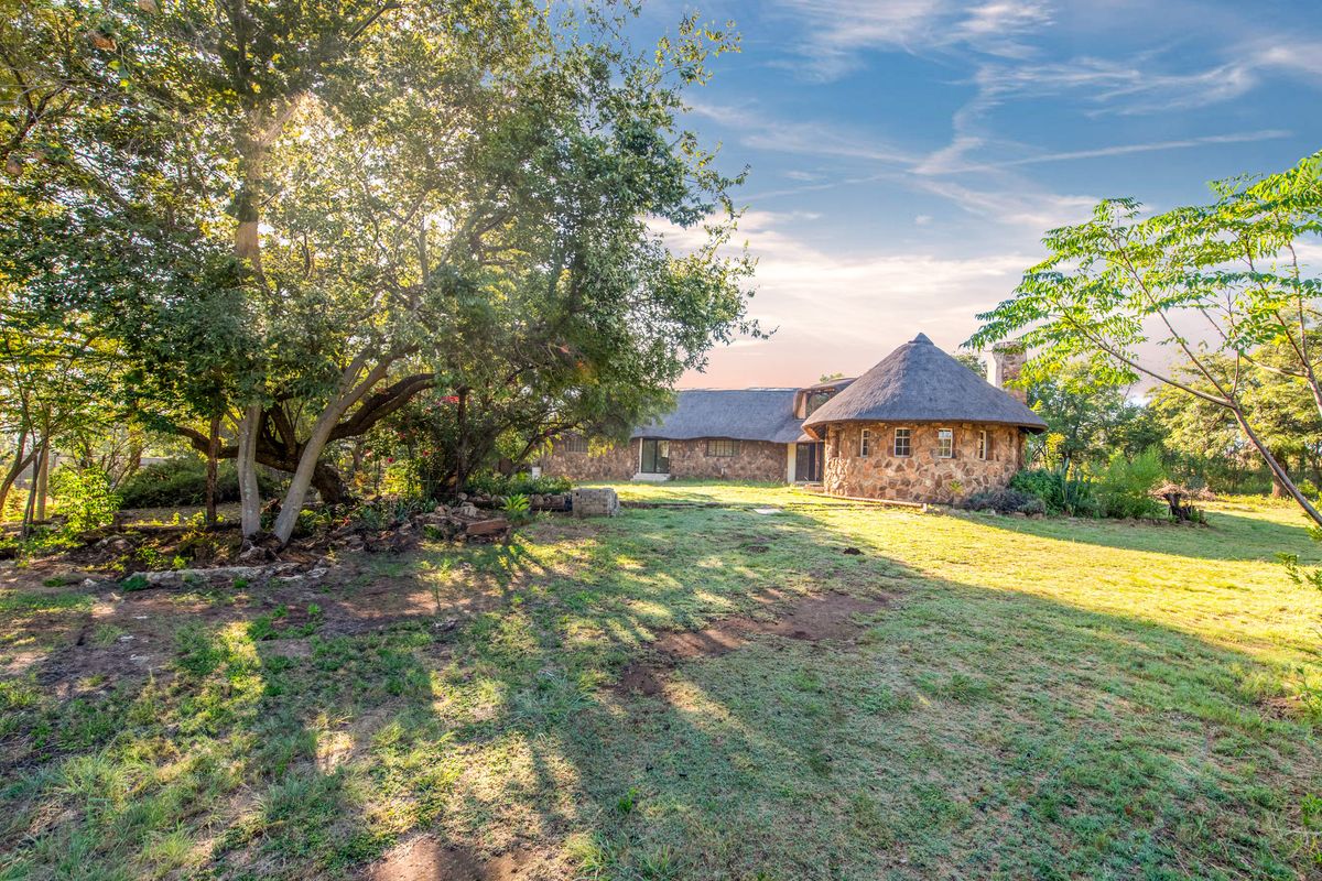 A scenic view of the rear elevation of a stone-clad home featuring a traditional thatched roof, set within a spacious, tree-lined garden.