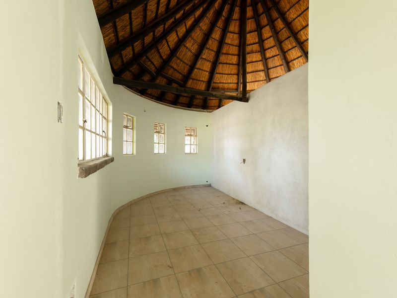 An empty, circular living area featuring a traditional thatched roof with exposed timber rafters, tiled flooring, and multiple steel-framed windows.
