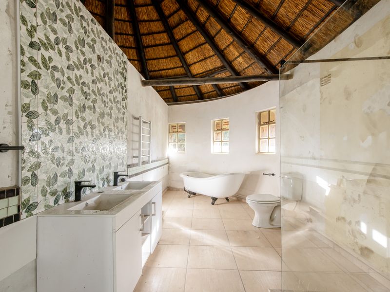 Spacious bathroom featuring a double vanity, freestanding bathtub, and glass shower, set under a traditional thatch roof with significant wall finish repairs required.