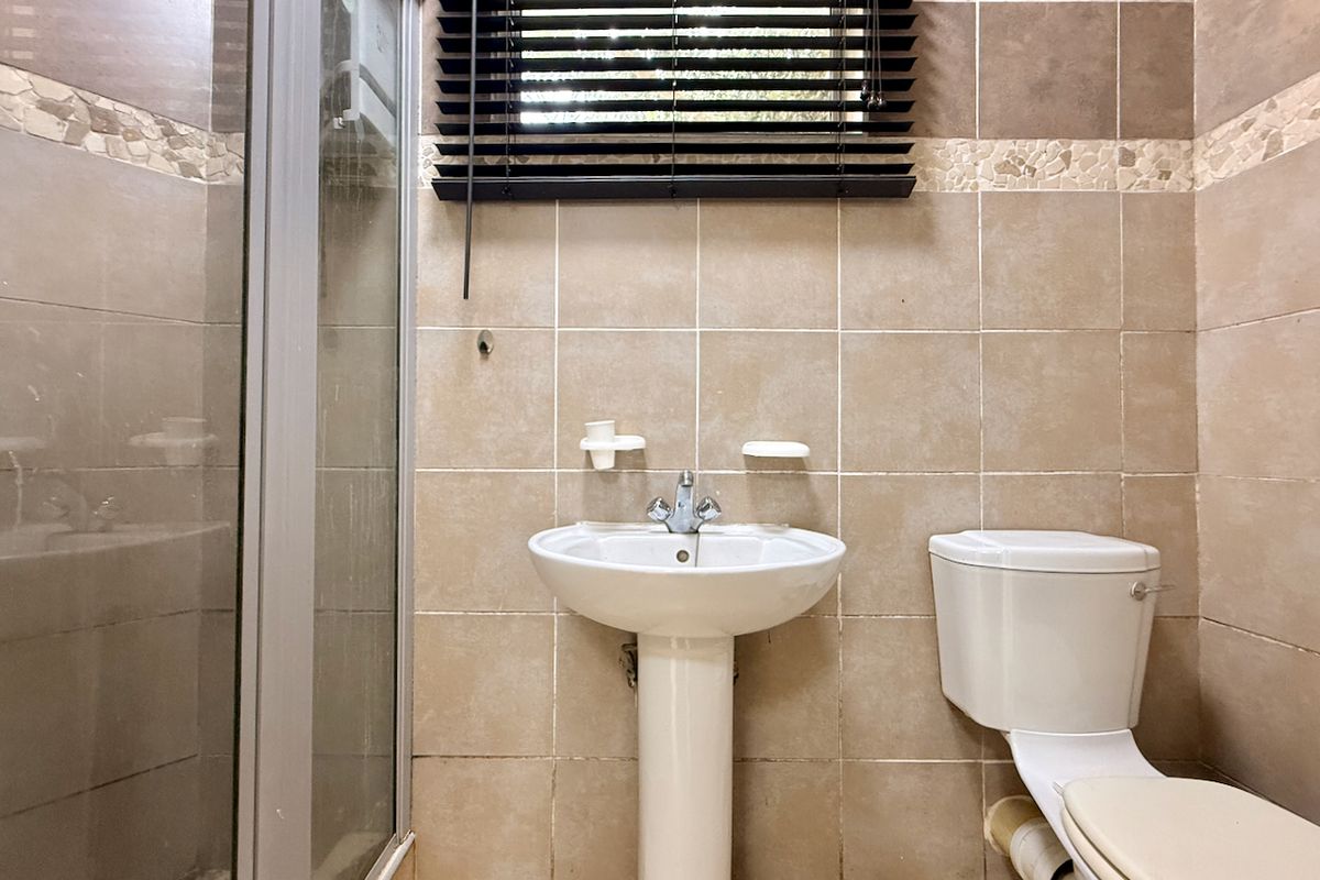 A functional bathroom featuring a pedestal basin, toilet, and shower enclosure with neutral ceramic wall tiling and venetian blinds.