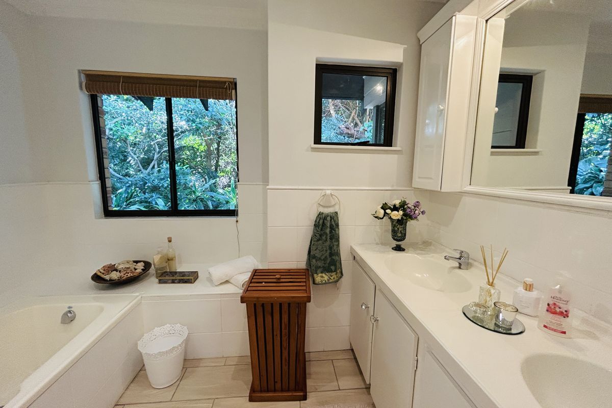 A bright bathroom featuring a built-in bathtub, double vanity with integrated mirror cabinetry, and large windows overlooking a lush garden.