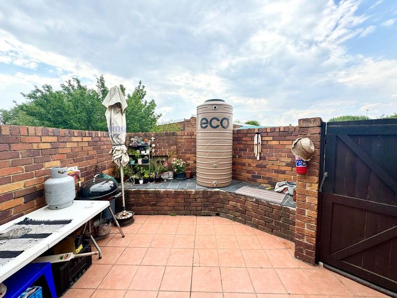 A paved outdoor patio area featuring a brick boundary wall, a water storage tank, and a timber access gate.