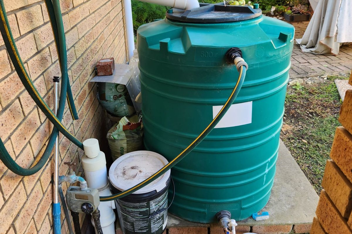 A functional exterior area featuring a 500-liter rainwater harvesting tank connected to the building's gutter system and an outdoor irrigation setup.