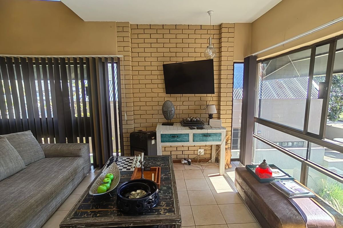 A living room featuring an exposed brick feature wall, tiled flooring, and large windows with vertical blinds, offering a bright and functional living space.