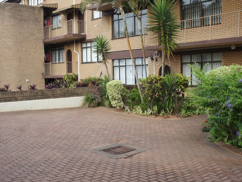 Front exterior view of a multi-story brick apartment building featuring paved driveway access, balconies, and established garden landscaping.
