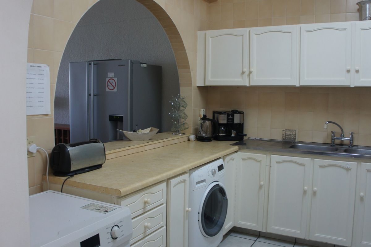A functional kitchen featuring white cabinetry, laminate countertops, a stainless steel sink, and a tiled backsplash, framed by a decorative archway.