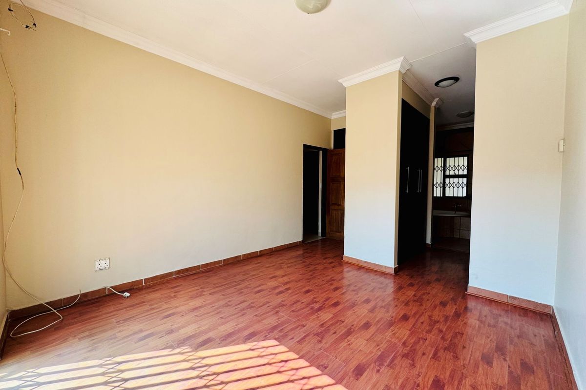 An empty living area featuring wood-look laminate flooring, crown molding, and built-in storage cupboards, with an adjacent hallway leading to a kitchen area.