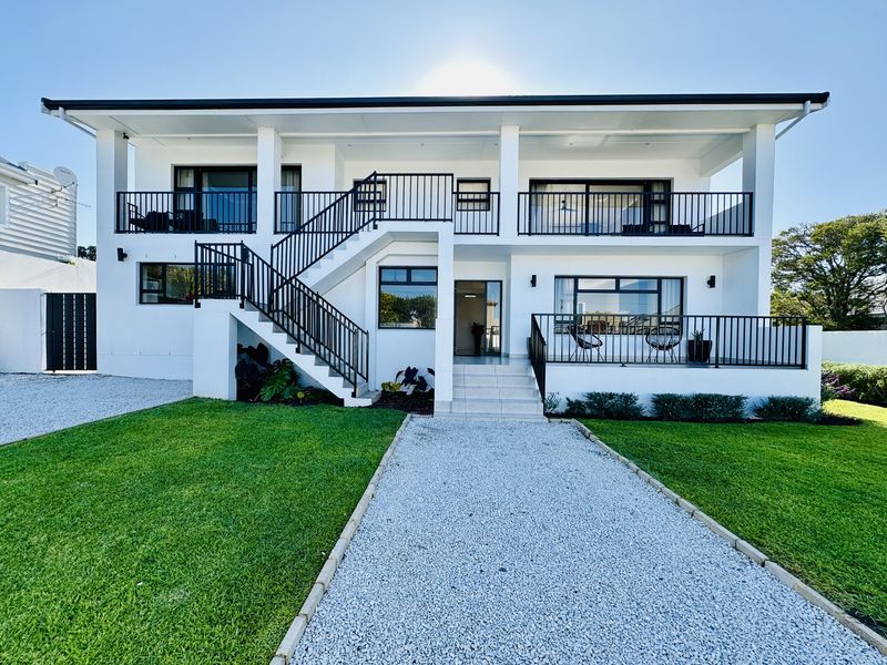 Modern two-story residential facade featuring a gravel driveway, manicured lawn, external staircase, and upper-level balcony with black metal railings.