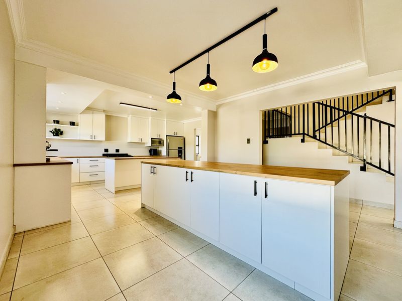 Spacious open-plan kitchen featuring a large island with a wooden countertop, modern cabinetry, tiled flooring, and an adjacent staircase with a metal railing.