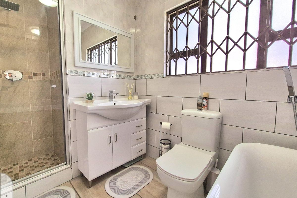 A functional bathroom featuring a shower enclosure, vanity basin, toilet, and bathtub with tiled finishes and security bars on the window.