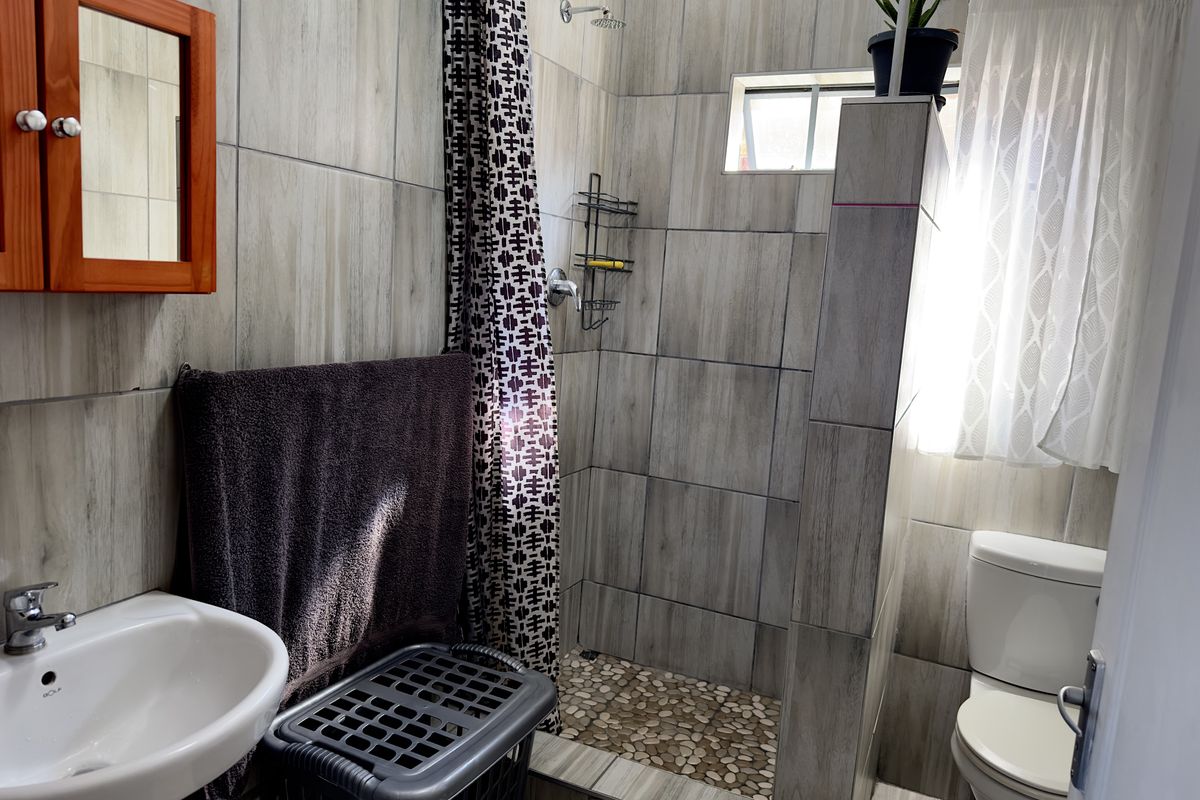 A compact bathroom featuring a walk-in shower with pebble-style flooring, wall-mounted basin, and a separate toilet area with neutral wood-look wall tiling.