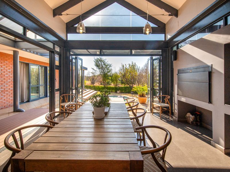 A bright, modern enclosed patio featuring a high vaulted ceiling, exposed steel beams, large glass folding doors, and a built-in fireplace.