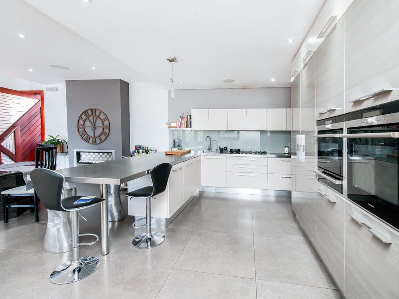 Modern open-plan kitchen featuring sleek cabinetry, integrated double ovens, a stainless steel breakfast bar, and large format floor tiles.