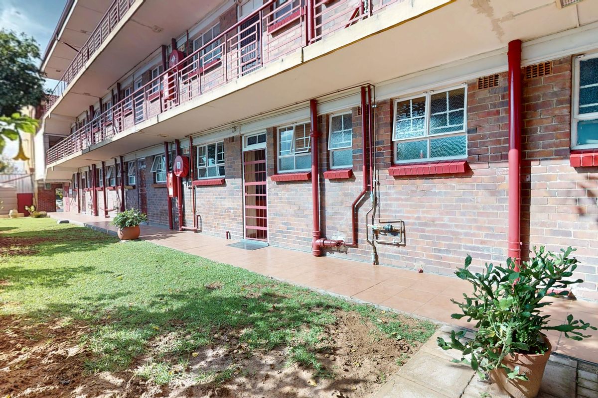 Rear exterior view of a multi-story brick apartment building featuring a tiled ground-level walkway, metal balcony railings, and external plumbing fixtures.