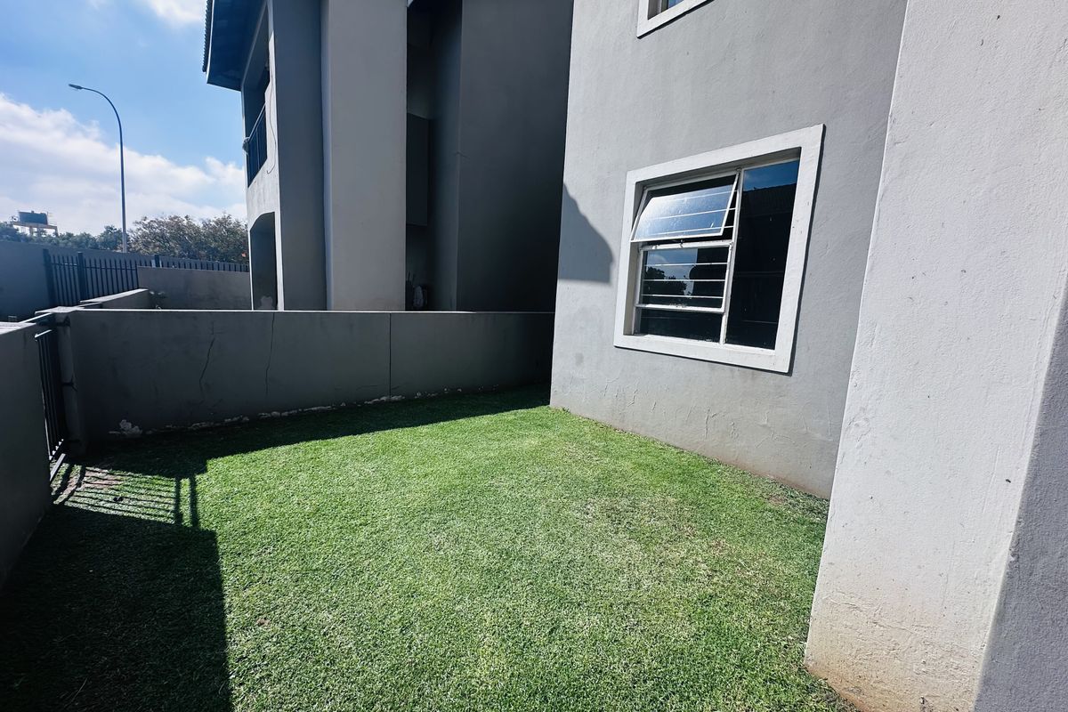 A private rear garden area featuring a lawn, enclosed by a boundary wall and metal fencing, adjacent to the apartment building exterior.